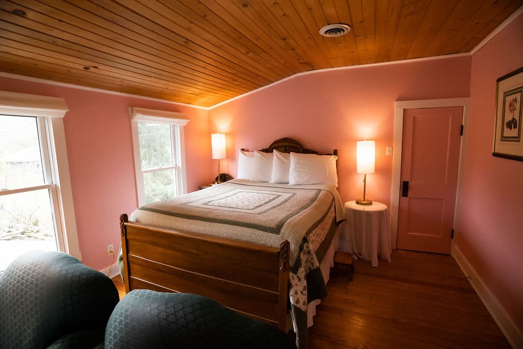 Queen size bed with four pillows, night stands with lamps atop and windows to garden