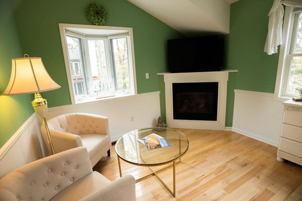 Sitting area by 3 windows with round table and two chairs, next to fireplace and flatscreen TV