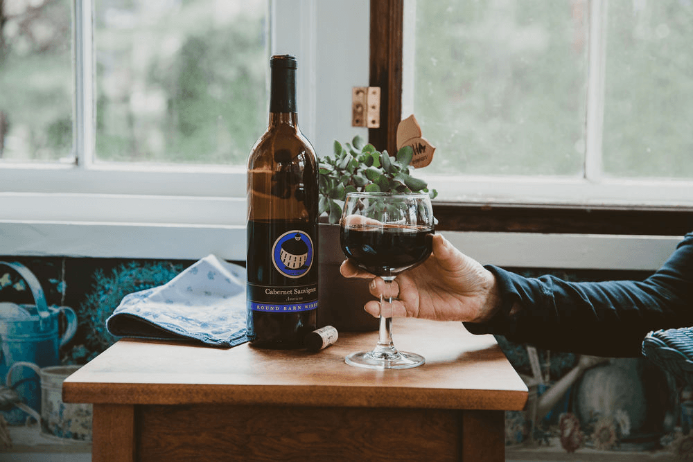 A hand holds a glass of red wine next to a bottle of Cabernet Sauvignon on a wooden table with a small plant and a napkin.