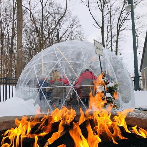 A transparent igloo with diners inside, surrounded by snow and a fire pit in front.