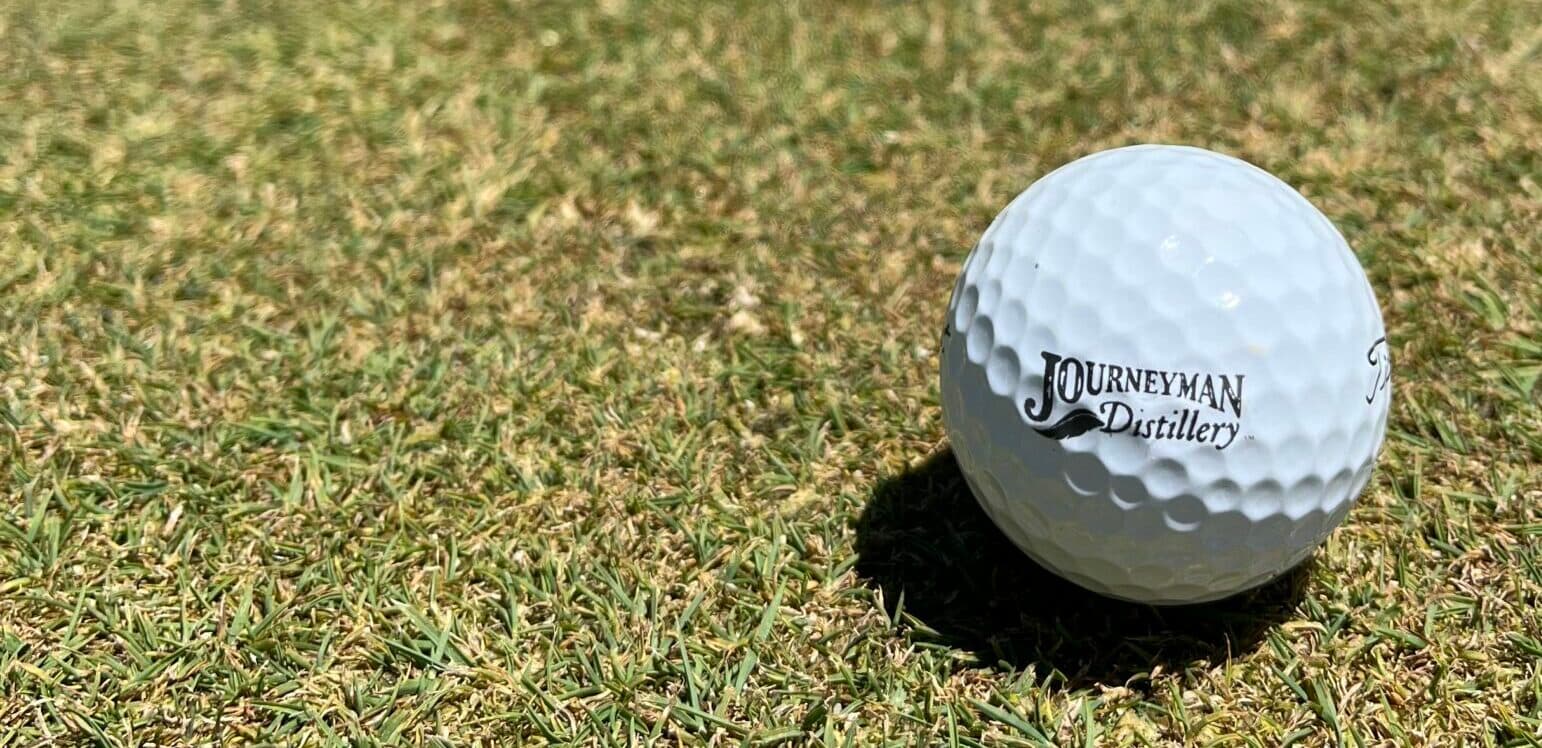 A white golf ball with "Journeyman Distillery" printed on it resting on green grass. A white golf ball with "Journeyman Distillery" printed on it resting on green grass.