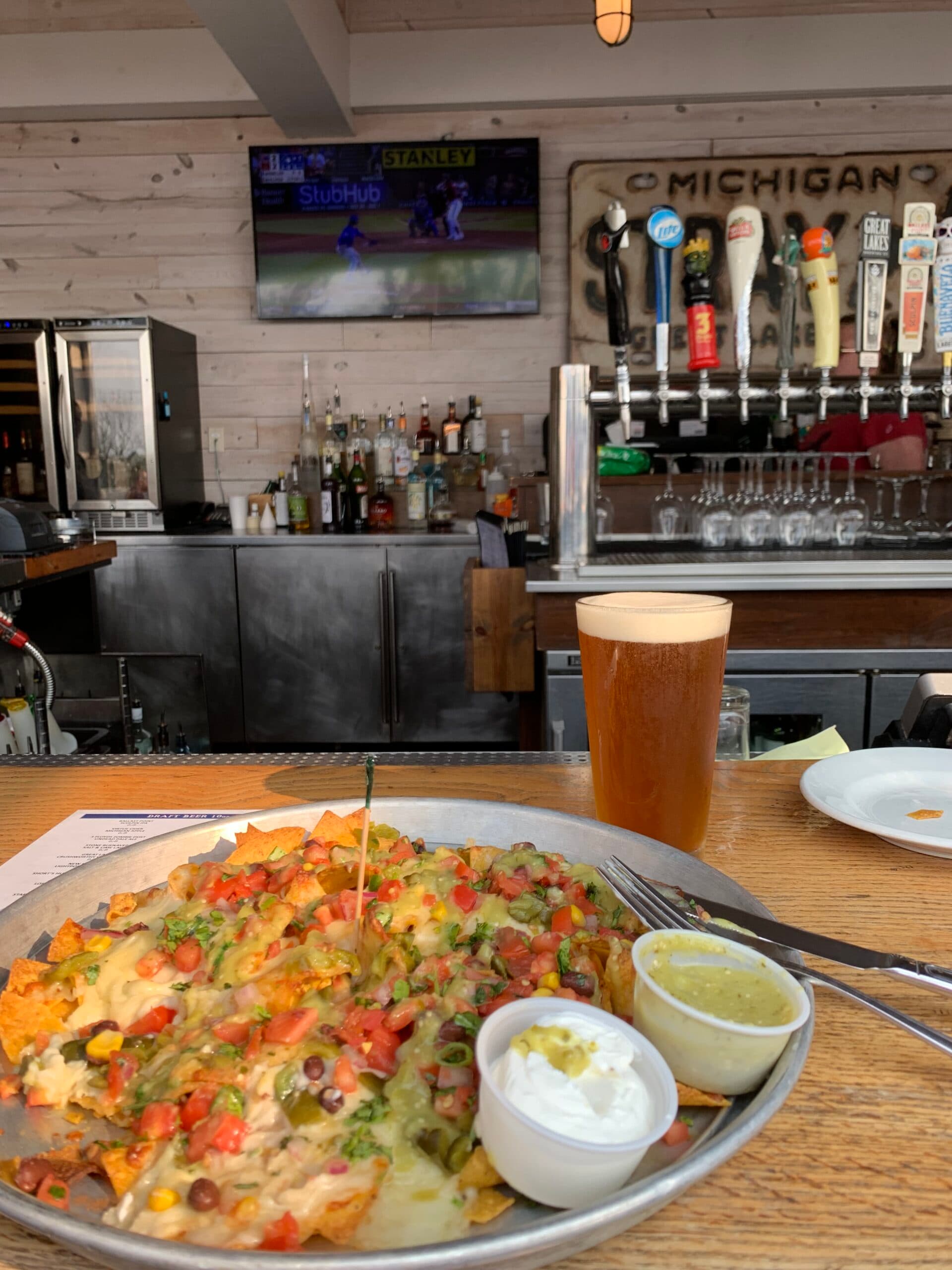 A plate of loaded nachos with toppings, accompanied by a pint of beer, sits on a bar counter with a television showing a sports game in the background.