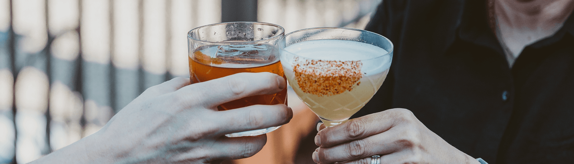 Two hands clink glasses holding a cocktail and a whiskey drink.