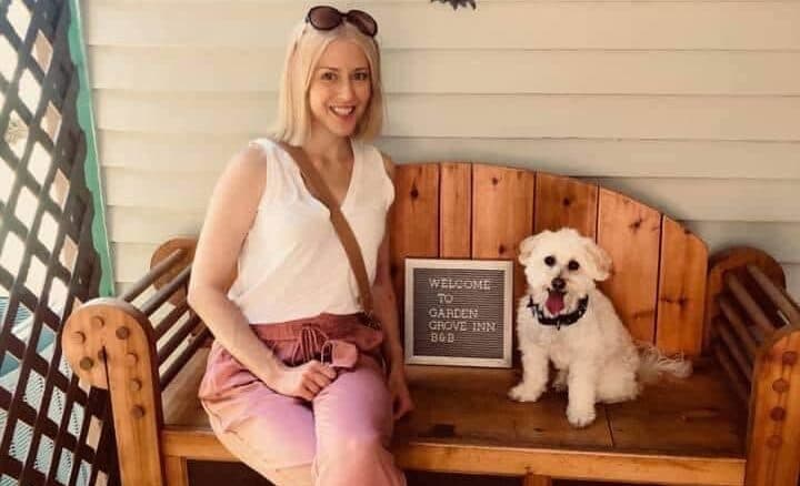 A woman sits on a wooden bench beside a small white dog, with a sign that says "Welcome to Garden Grove Inn." A woman sits on a wooden bench beside a small white dog, with a sign that says "Welcome to Garden Grove Inn."