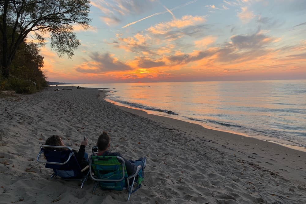 Two people sitting in beach chairs on a sandy shore watching a colorful sunset over the water.