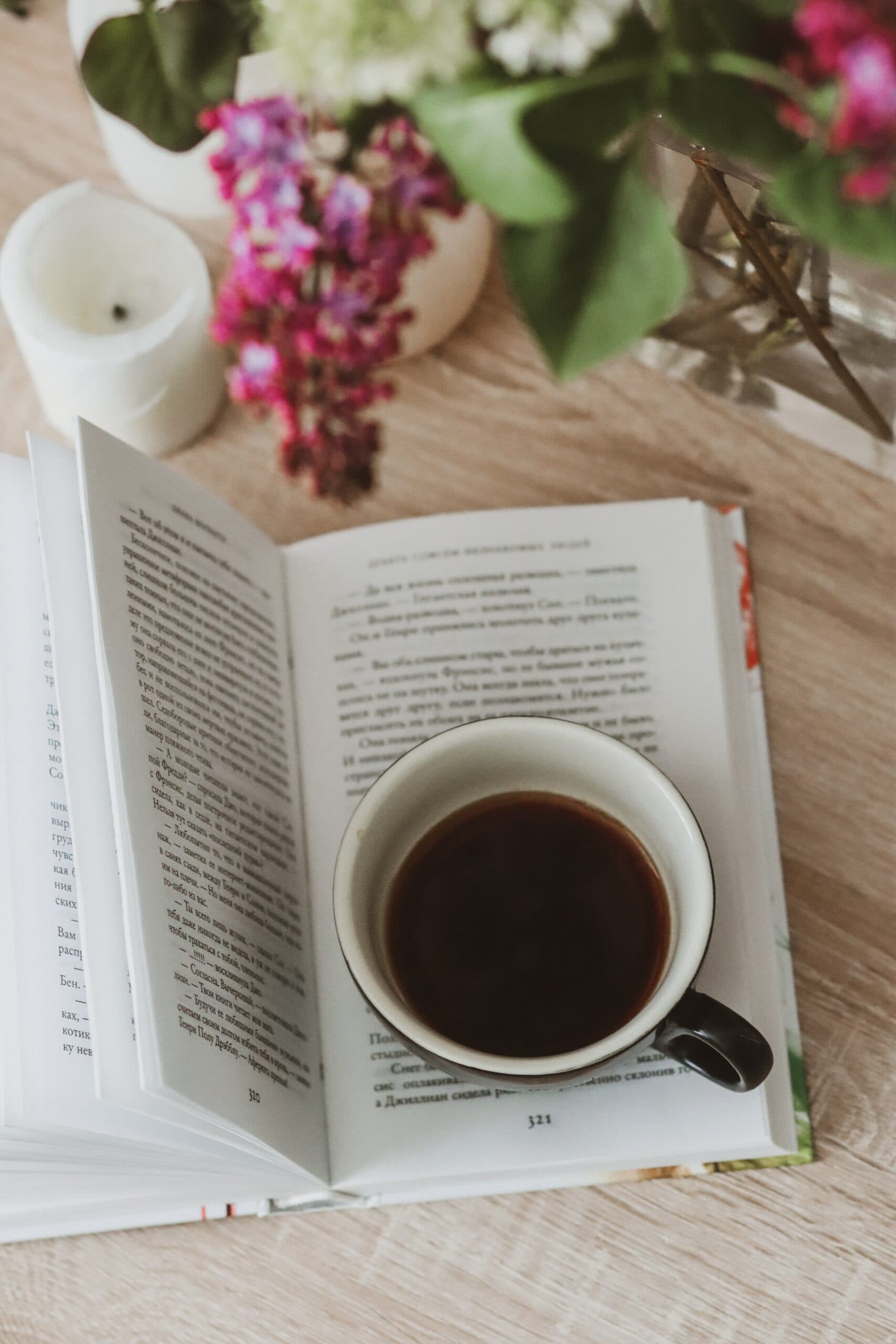 A cup of coffee rests on an open book, surrounded by flowers and a candle. A cup of coffee rests on an open book, surrounded by flowers and a candle.