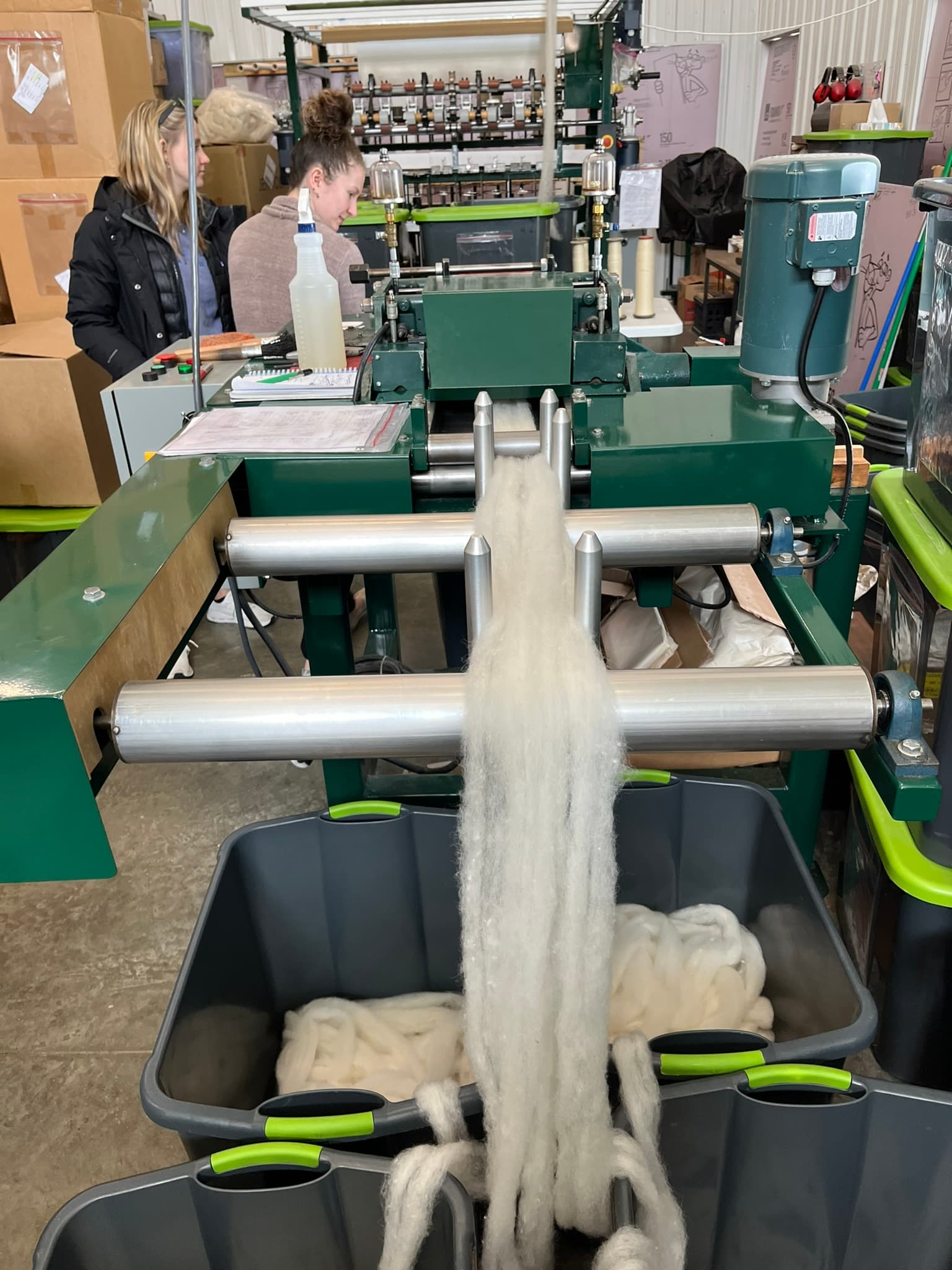 A wool processing machine dispenses wool into containers while two women work nearby.