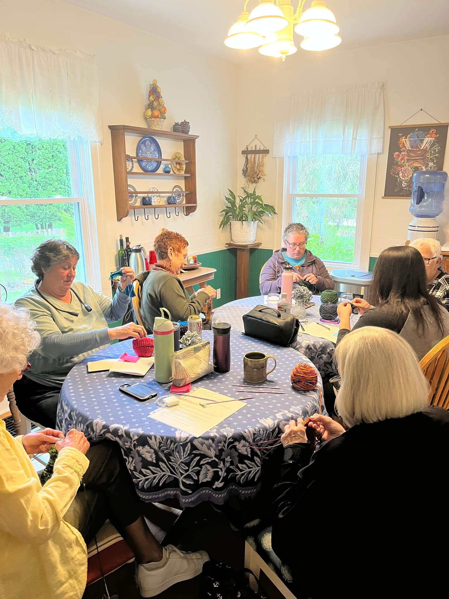 A group of six people sit around a table, engaged in a crafting activity in a cozy room filled with natural light.