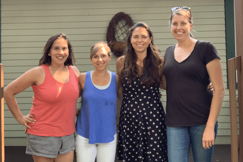 Four women stand together outside, smiling in casual attire.