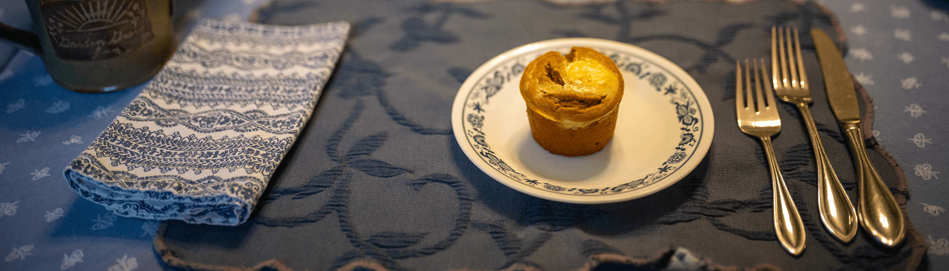 A plate with a cupcake is arranged on a patterned tablecloth next to folded napkins and cutlery.