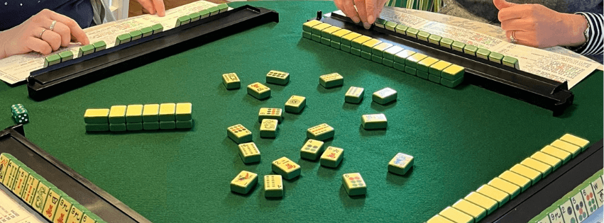 Two players' hands are visible as they play a game of Mahjong on a green table. Two players' hands are visible as they play a game of Mahjong on a green table.
