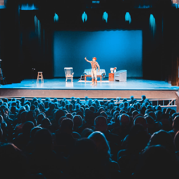 A performer gestures on stage in front of a seated audience, with a simple set of furniture in the background.