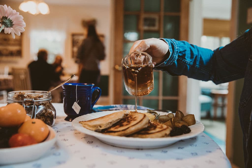 A hand pours tea over a plate of pancakes at a cozy café table, accompanied by a blue mug and a bowl of fresh fruit.