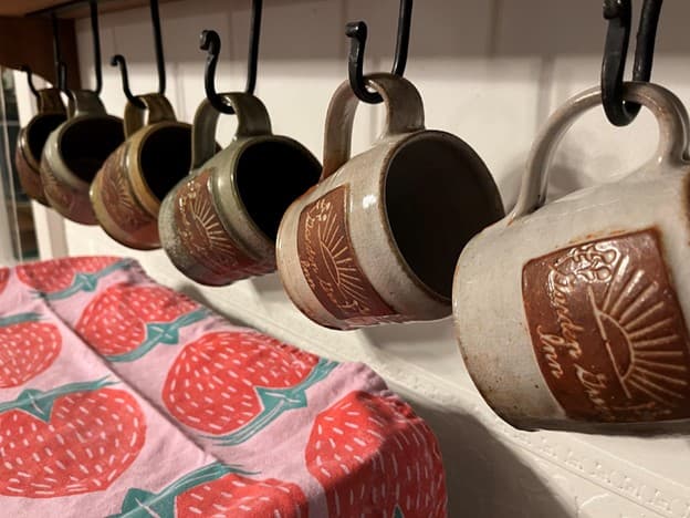 A row of ceramic mugs hanging from hooks above a strawberry-patterned towel.