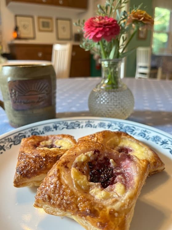 A plate of three pastry tarts with fruit filling, accompanied by a decorative vase of flowers and a mug, in a cozy kitchen setting.