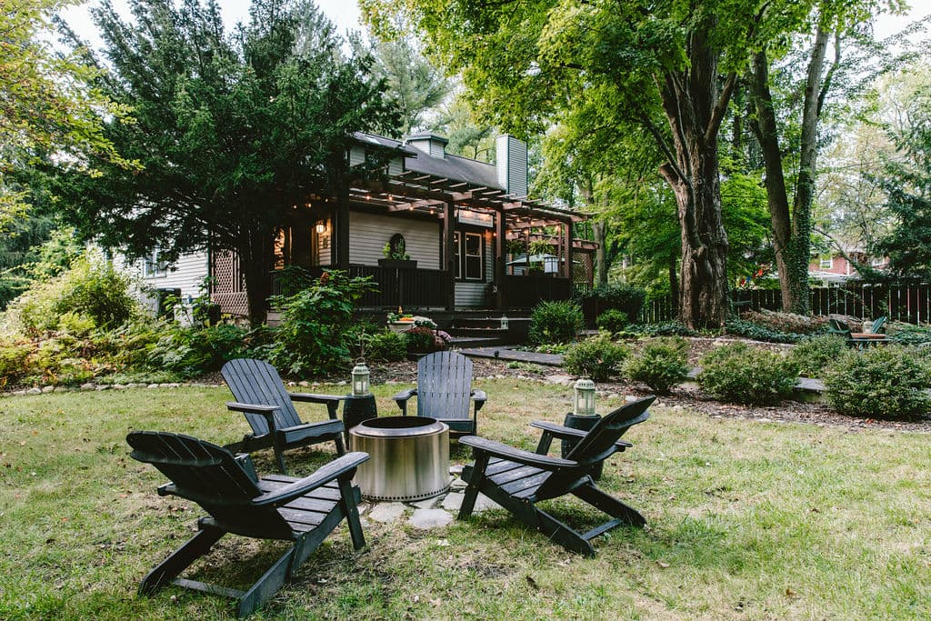 A cozy backyard with four Adirondack chairs arranged around a fire pit, surrounded by lush greenery and a house in the background. A cozy backyard with four Adirondack chairs arranged around a fire pit, surrounded by lush greenery and a house in the background.