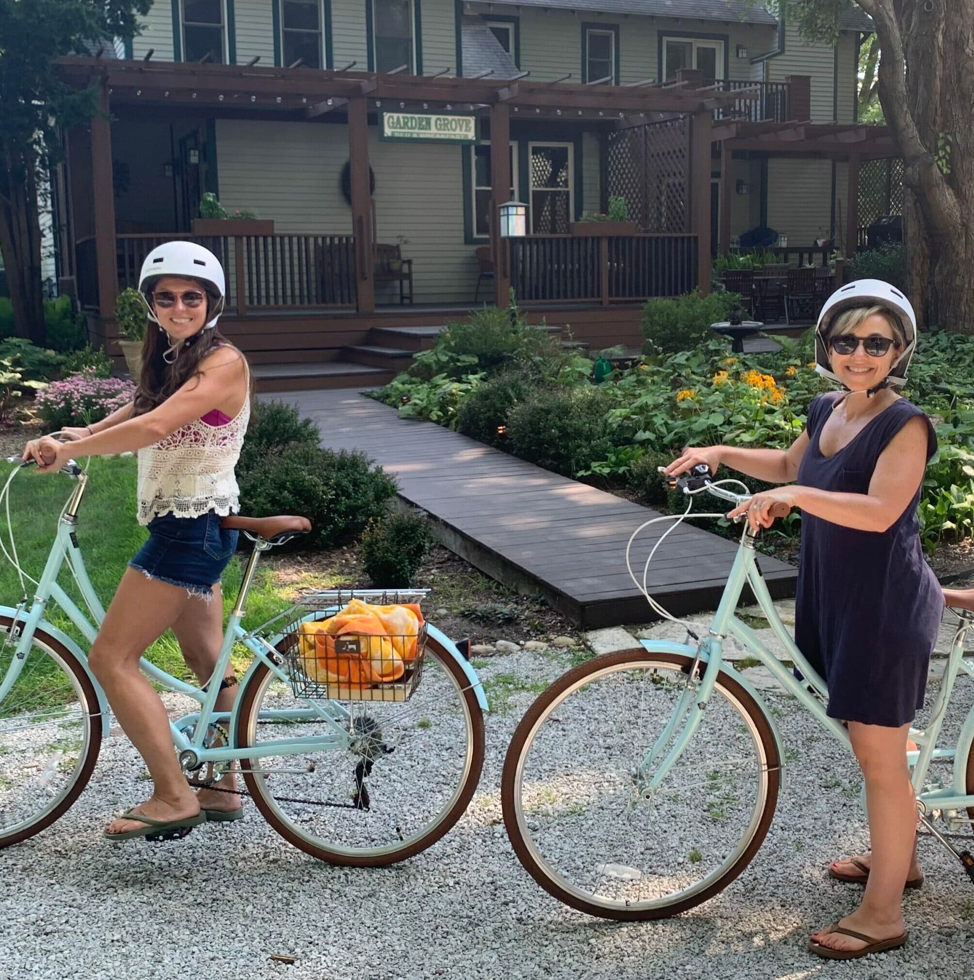 Two women wearing helmets pose on pastel bicycles in front of a house with a garden.