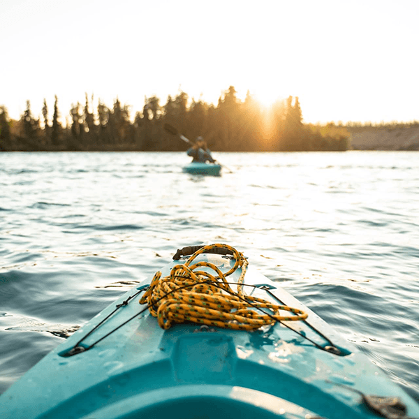 A close-up of a blue kayak with a coiled yellow rope, with another kayaker paddling in the background under a sunset.