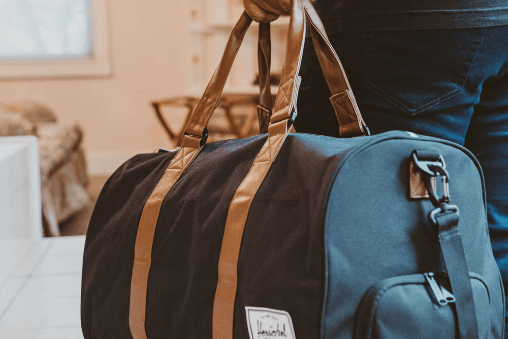 A person holds a dark duffel bag with brown straps in an indoor setting.