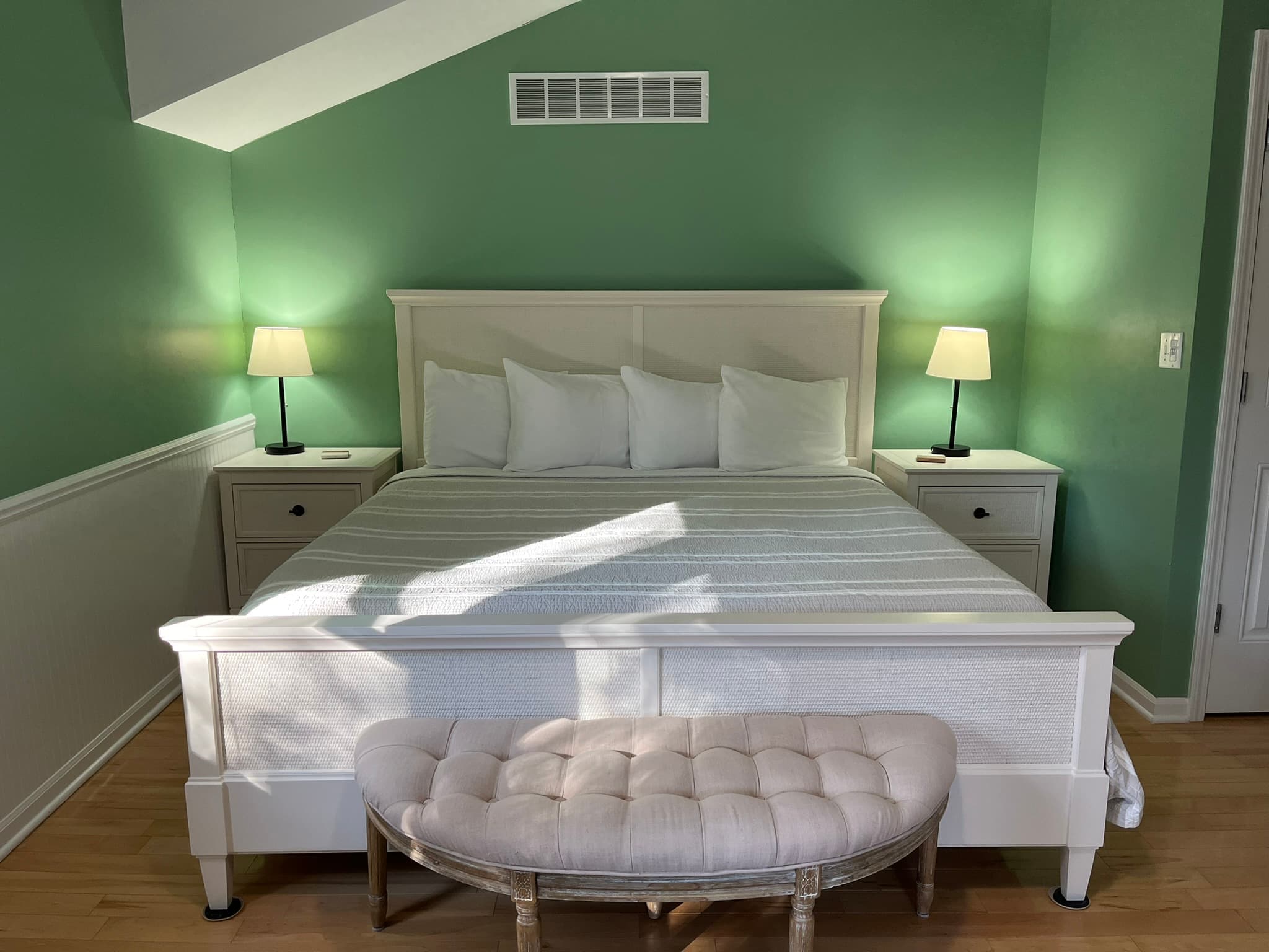 A neatly made bed with white linens features two bedside lamps in a green-walled bedroom.