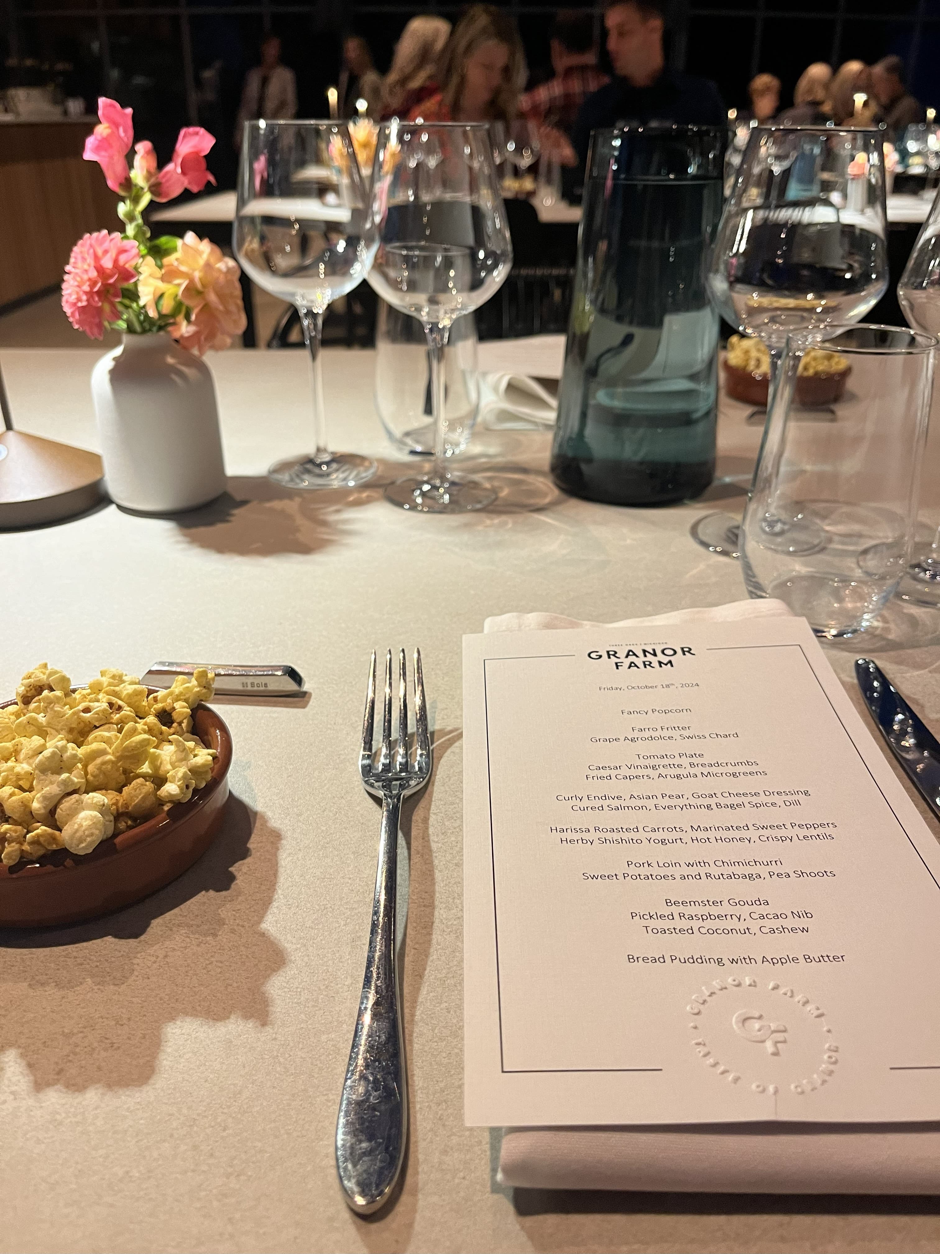 A dining table setting featuring a menu, fork, bowl of popcorn, flowers, and wine glasses. A dining table setting featuring a menu, fork, bowl of popcorn, flowers, and wine glasses.