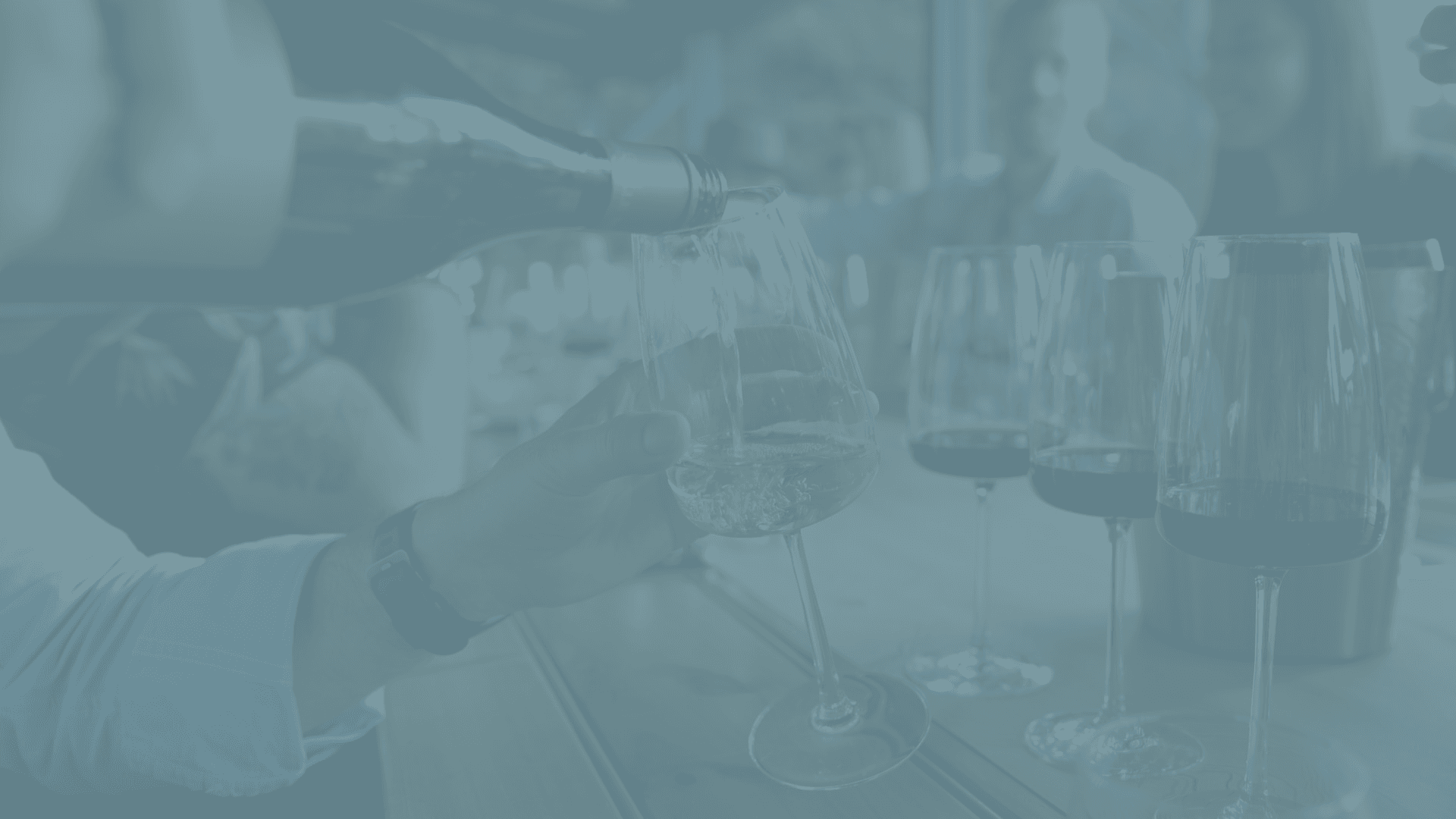 A hand pours white wine into a glass at a social gathering with blurred people in the background.