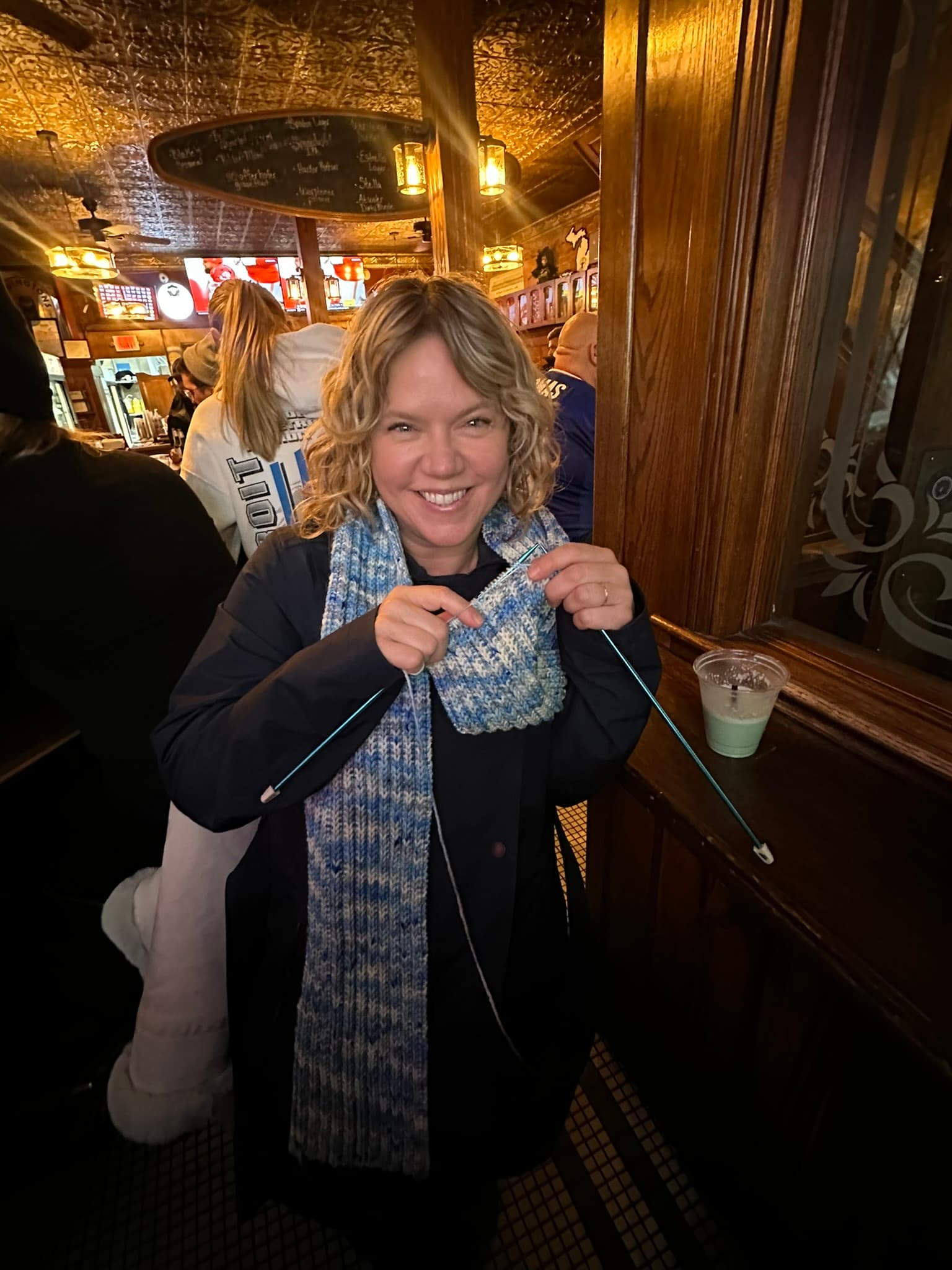 A smiling woman in a black coat holds knitting needles in a cozy pub setting.